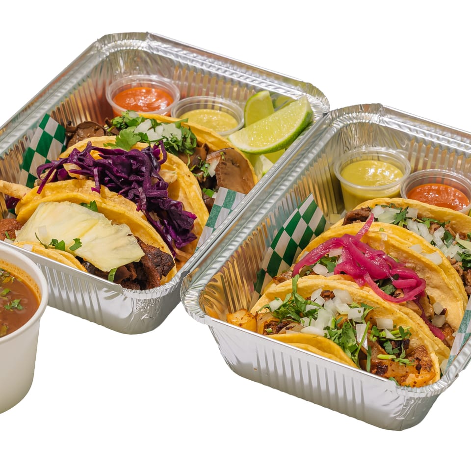 Two trays of tacos topped with cilantro, onions, cabbage, pineapple, and pickled vegetables, served with lime wedges, sauces, and consommé.